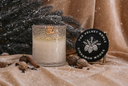 A lit scented candle in a textured clear glass jar sits on a beige fabric surface beside a round black decorative lid reading 'THE VELVET NOBLE CINNAMON WOODS.' The scene is styled with pine branches, cinnamon sticks, dried botanicals, and falling snowflake effects, creating a warm holiday atmosphere.