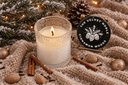 A lit white candle in a textured glass holder sits on a beige knit blanket with cinnamon sticks, walnuts, pinecones, and holiday lights. A round black decorative lid reading 'THE VELVET NOBLE CINNAMON WOODS' is visible on the right.