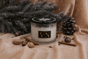 A glass jar candle with a black lid and a black label reading 'VN' in gold lettering, styled on a beige fabric surface with cinnamon sticks, nuts, pinecones, and dark evergreen branches, with a snowy overlay effect.