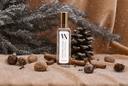 A clear glass bottle of Cinnamon Woods Air & Linen Mist with a gold cap and white label, styled on a beige fabric surface with cinnamon sticks, pinecones, and snow-dusted evergreen branches in a warm, festive setting.