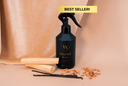Black spray bottle labeled 'Velvet Home' with wooden sticks and vanilla beans on a beige fabric background