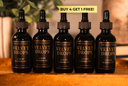 Five black dropper bottles labeled 'The Velvet Noble Velvet Drops Luxury Warmer Oil' arranged on a wooden surface with a blurred indoor background. A yellow banner at the top reads 'BUY 4 GET 1 FREE!'.