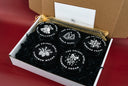 An open white gift box filled with black shredded paper and five round black tins labeled 'The Velvet Node' with different scented notes, set against a dark red background.
