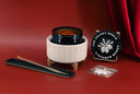 A candle product set on a red background, featuring a cream ribbed jar with a black lid, a black branded candle tin reading 'The Velvet Noble Cinnamon Woods,' a long black scented stick, and a small card.