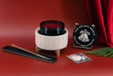 A festive holiday candle set displayed on a red surface with a red curtain backdrop. The set includes a white ceramic candle warmer with a black lid and red wax, a black decorative plaque reading 'THE VELVET NOBLE VELVET HOLLYDAYS', a long black tray with a gold wand, a small card, and green pine branches.