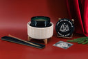 A festive Christmas-themed product set on a red background, featuring a cream ribbed ceramic holder with a dark green glass vessel, a black circular plaque reading 'THE VELVET NOBLE NOBLE NOEL' with a white tree design, a long black tray with a gold-toned rod, and a small printed card, styled with red draped fabric and green pine branches.