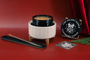 A festive Christmas-themed product set on a red background, featuring a cream ribbed ceramic holder with a dark green glass vessel, a black circular plaque reading 'THE VELVET NOBLE Cashmere Cedar' with a white tree design, a long black tray with a gold-toned rod, and a small printed card, styled with red draped fabric and green pine branches.