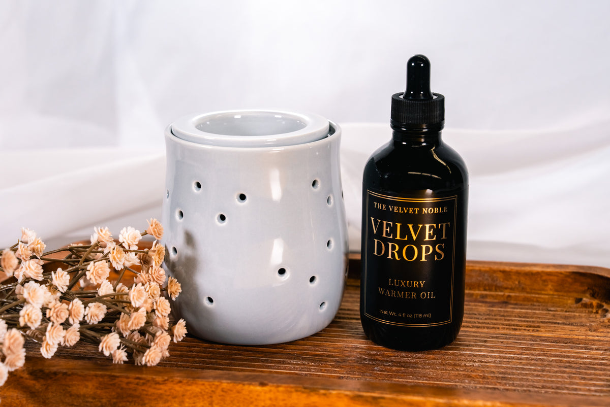 Bottle of 'The Velvet Noble Velvet Drops' oil on a wooden surface with a light blue ceramic warmer and dried flowers.