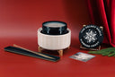 A holiday-themed candle set on a red background, featuring a cream ribbed candle holder with a black lid, a long black and gold taper candle, a round black tin labeled 'THE VELVET NOBLE DIAMOND FROST', and a snowflake card with pine branches.