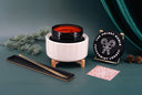 A styled product shot of a candle in a ribbed white ceramic pot with a black lid, set on a dark teal surface with holiday greenery, pinecones, a black sign reading 'The Velvet Noble Peppermint Legacy,' and a small patterned card.
