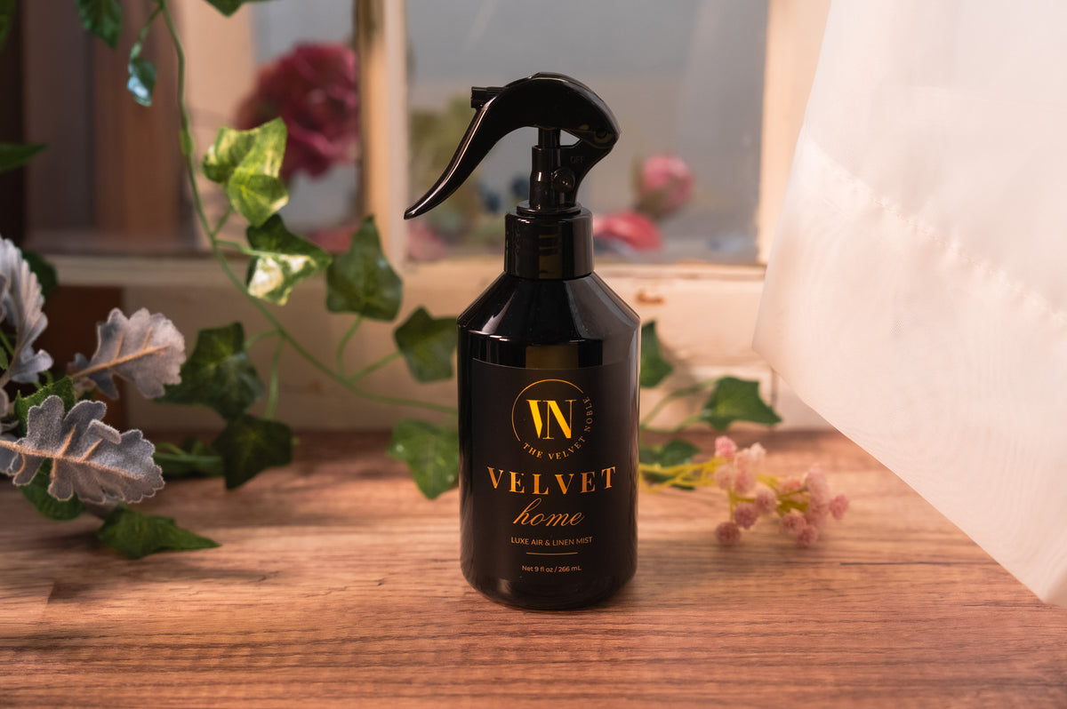 A bottle of Velvet Home on a wooden surface with flowers in the background.