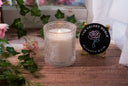 A single wick blushed rain candle on a wet wooden surface surrounded by flowers and greenery