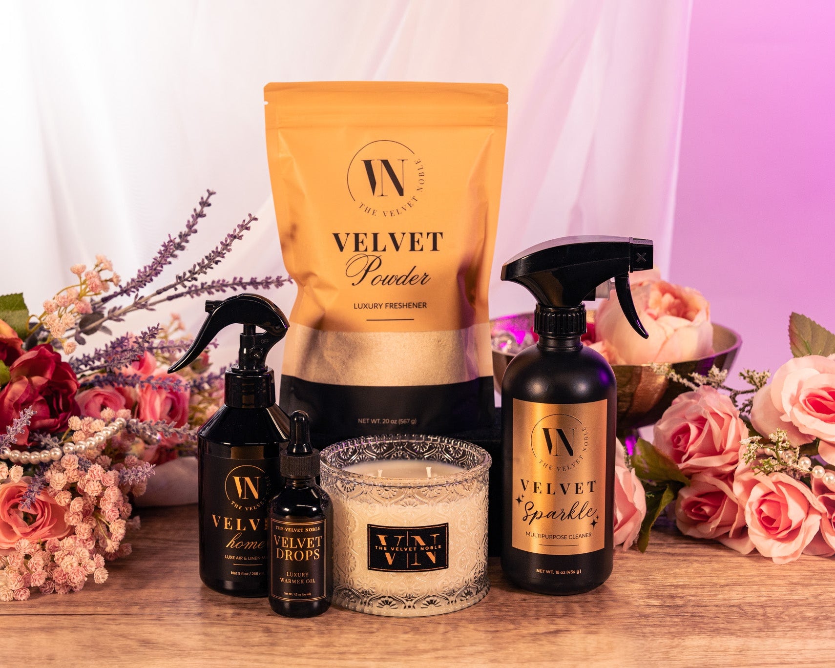 Collection of 'Velvet' brand products including a bag, bottles, and a candle on a table with flowers in the background.