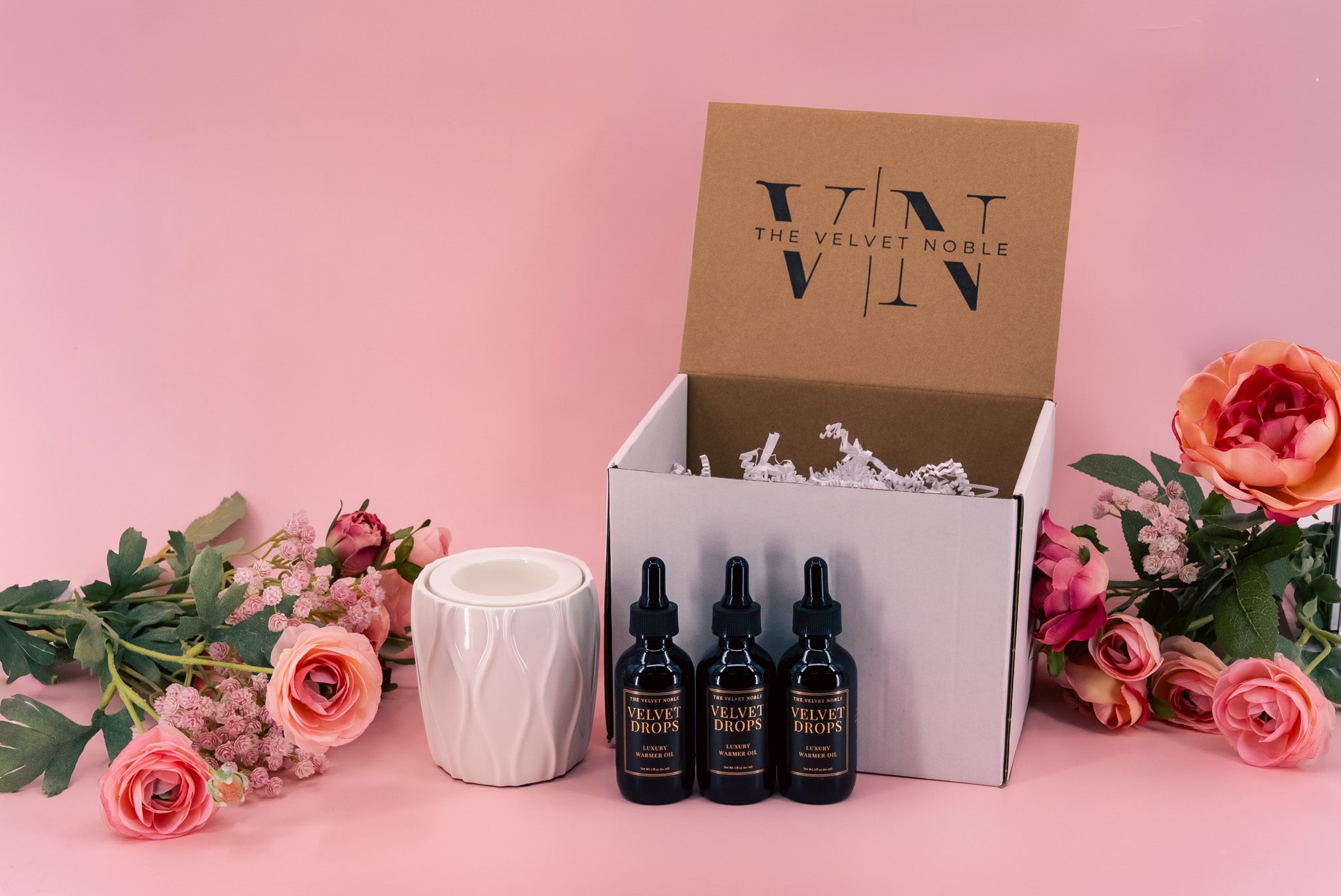 3 bottles of velvet drops with a white petite warmer and Velvet Noble packaging on a pink background with flowers.