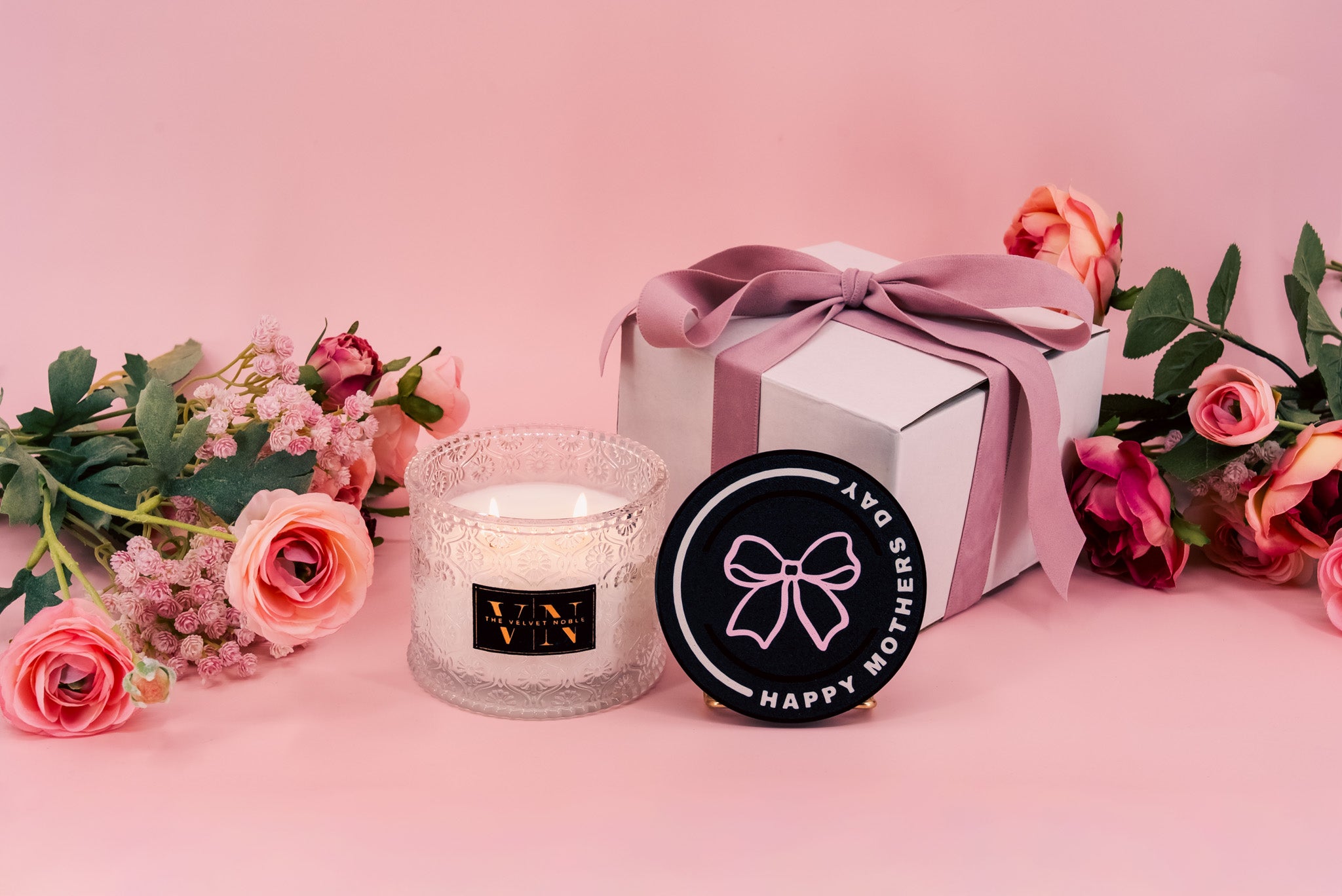 Candle, mother's day candle lid, & gift box with pink ribbon, and flowers on a pink background