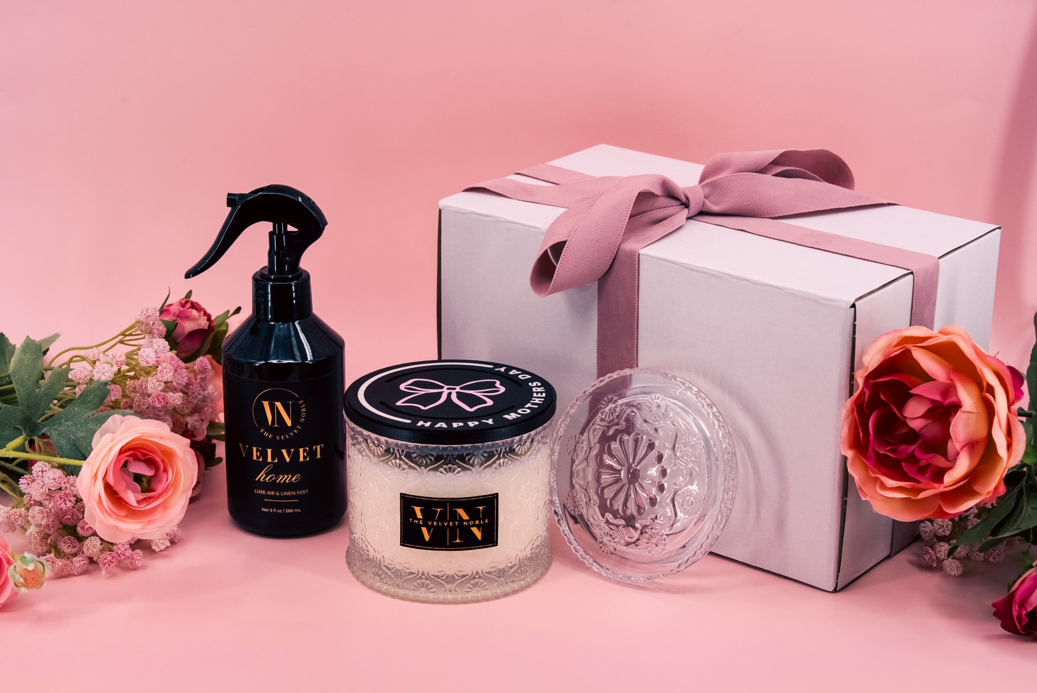Velvet Noble 2-wick candle with a mother's day lid and glass lid, bottle of Velvet Home, and gift packaging on a pink background with flowers.