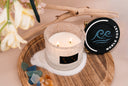 A lit Noble Coast 2 wick candle on a wood tray with sand, sea glass, amber and the lid on the side.