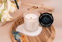 Noble Coast single wick candle atop a pile of sand on a wooden tray. There is also driftwood, florals, sea glass, and amber