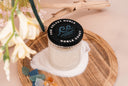 Noble Coast single wick candle atop a pile of sand on a wooden tray. There is also driftwood, florals, sea glass, and amber