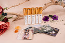 A set of 5 small linen mist bottles with golden caps. The labels say Velvet Ease, Blushed Rain, Washed Amber, Morning Mist, and Black Vintage. Surrounded by flowers, amber, driftwood, flowers and 5 cards for each individual fragrance.