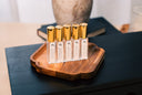 A set of 5 small linen mist bottles with golden caps. The labels say Velvet Ease, Blushed Rain, Washed Amber, Morning Mist, and Black Vintage. All on a wooden tray.