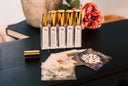 A set of 5 small linen mist bottles with golden caps. The labels say Velvet Ease, Blushed Rain, Washed Amber, Morning Mist, and Black Vintage. All on a black wooden surface.