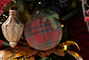 A close-up of a red and green frosted plaid Christmas ornament hanging on a decorated Christmas tree, next to a textured metallic gold ornament.