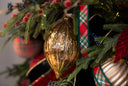 A close-up shot of a Gold Teardrop Ornament that's on a Christmas tree branch with green pine needles, small red berries, a shiny gold ornament, red plaid ribbon, and other holiday decorations.