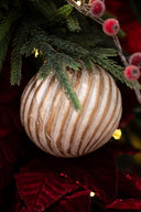 A close-up of a round Christmas ornament with vertical beige and white stripes hanging among green pine branches and red holiday decorations.