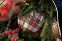 A close-up of a round Christmas ornament with a red, blue, and white plaid pattern, nestled among green pine branches with small warm lights and red berries.