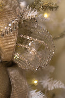 A close-up of a clear glass ornament decorated with silver rhinestone swirls, hanging on a white artificial Christmas tree with gold tinsel and a beige ribbon.