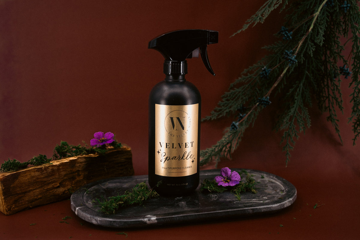 Black spray bottle labeled 'Velvet Sparkle' on a dark surface with greenery and flowers.