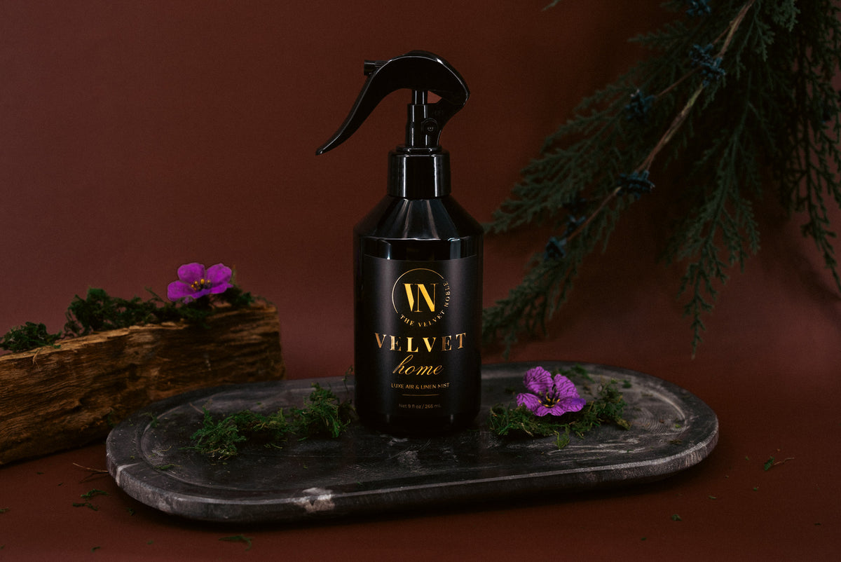 Black spray bottle labeled 'Velvet Home' on a dark surface with purple flowers and greenery.