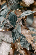 Close-up of decorative artificial foliage with dark green leaf-shaped pieces and glittery silver branches layered over a soft beige background.