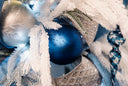 A large blue mercury glass round ornament, with a dotted finish, on a white and silver Christmas tree.