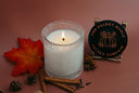 A lit white candle in a textured clear glass holder sits on a dusty rose background, styled with a red autumn leaf, cinnamon sticks, pinecones, and a round black label that reads 'THE VELVET NOBLE VELVET PUMPKIN'.