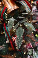 Close-up of a decorated Christmas tree branch with green artificial leaves, red and green ribbons, warm string lights, and a metallic gold ornament.
