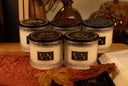 Five scented candles in textured glass jars with black lids, arranged on a wooden surface with autumn-themed props including a quilt, leaves, and pumpkins in the background. The front labels read THE VELVET NOBLE