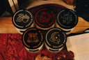 Five scented candles in textured glass jars with black lids, arranged on a wooden surface with autumn-themed props including a quilt, leaves, and pumpkins in the background. The front labels read THE VELVET NOBLE