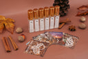 Six clear glass bottles with metallic gold caps and white labels are arranged on a dusty rose background with autumn-themed props including cinnamon sticks, acorns, a pinecone, dried leaves, and patterned scent cards.
