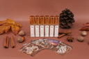 Six clear glass bottles with metallic gold caps and white labels are arranged on a dusty rose background, surrounded by autumn-themed props including a pinecone, cinnamon sticks, star anise, acorns, and printed scent cards.
