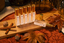 A set of clear glass bottles with gold caps and white labels, arranged on a wooden board with autumn leaves, cinnamon sticks, and acorns on a rust-colored fabric surface.