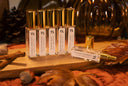 A set of clear glass bottles with gold caps and white labels, arranged on a wooden board with autumn leaves, cinnamon sticks, and acorns on a rust-colored fabric surface.