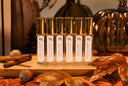 A set of clear glass bottles with gold caps and white labels, arranged on a wooden board with autumn leaves, cinnamon sticks, and acorns on a rust-colored fabric surface.