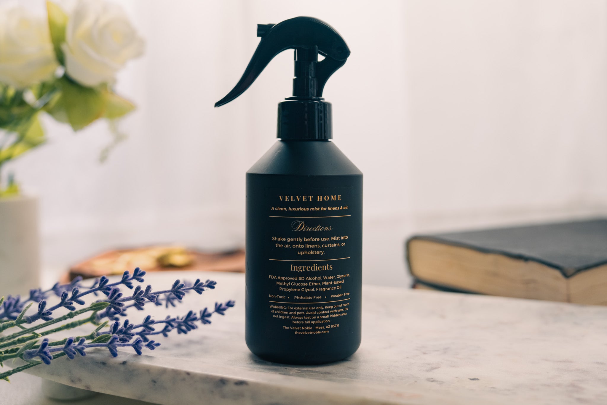 Black spray bottle labeled 'Velvet Home' on a marble surface with lavender and flowers in the background