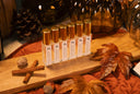 A set of clear glass bottles with gold caps and white labels, arranged on a wooden board with autumn leaves, cinnamon sticks, and acorns on a rust-colored fabric surface.