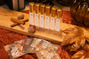 A set of clear glass bottles with gold caps and white labels, arranged on a wooden board with autumn leaves, cinnamon sticks, and acorns on a rust-colored fabric surface.