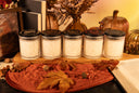 Five small jars of scented candles with black lids are arranged on a wooden board with autumn-themed props including dried leaves, books, pinecones, and decorative pumpkins. The lids show different designs and text including 'The Velvet Noble' and scent names like 'Vanilla Harvest' and 'Golden Twilight'.