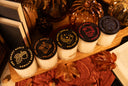 Five small jars of scented candles with black lids are arranged on a wooden board with autumn-themed props including dried leaves, books, pinecones, and decorative pumpkins. The lids show different designs and text including 'The Velvet Noble' and scent names like 'Vanilla Harvest' and 'Golden Twilight'.