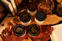 Five small jars of scented candles with black lids are arranged on a wooden board with autumn-themed props including dried leaves, books, pinecones, and decorative pumpkins. The lids show different designs and text including 'The Velvet Noble' and scent names like 'Vanilla Harvest' and 'Golden Twilight'.